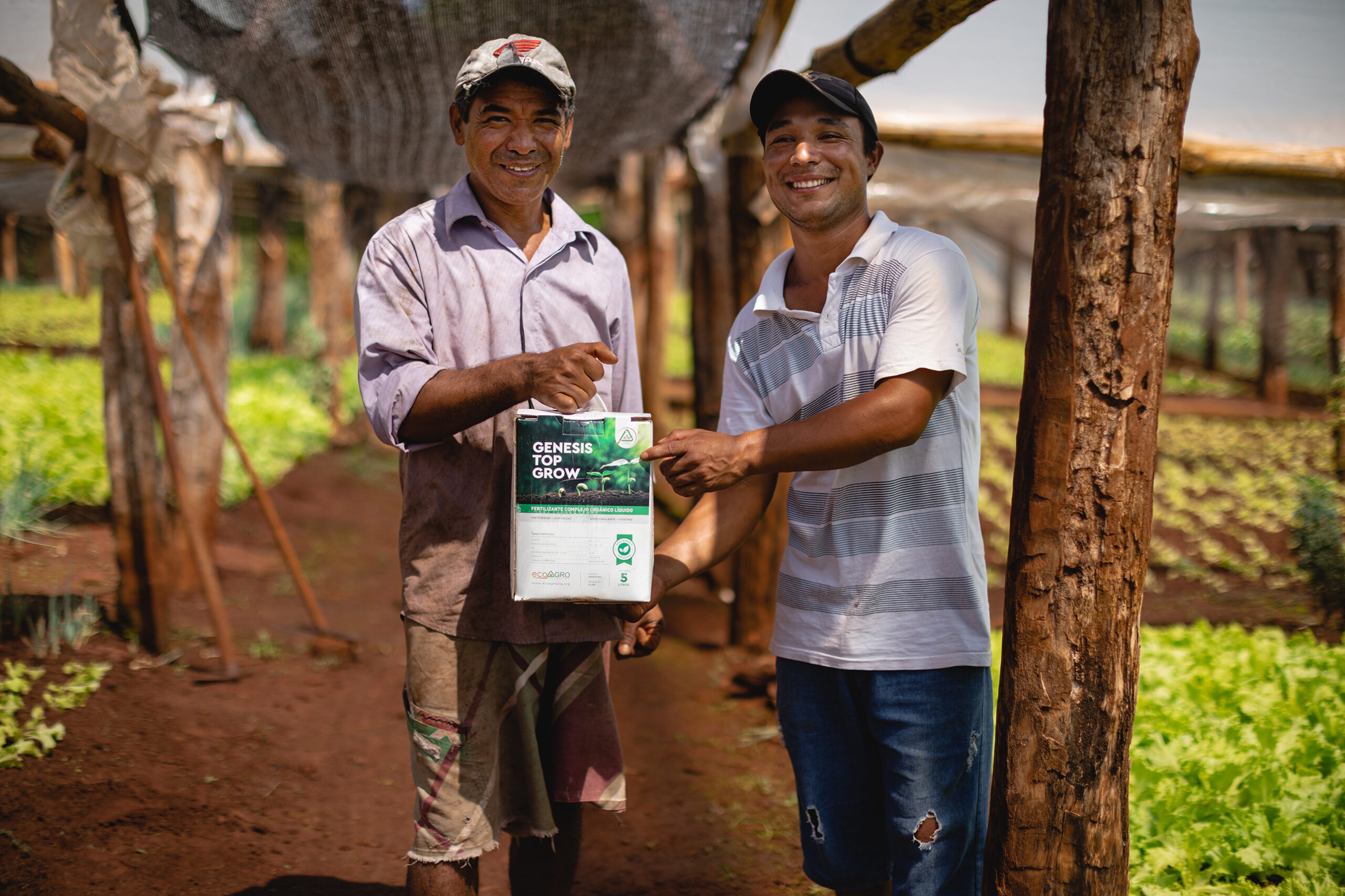 Dos agricultores sonrientes muestran un envase de biofertilizante natural en una huerta bajo cobertura, rodeados de cultivos verdes.