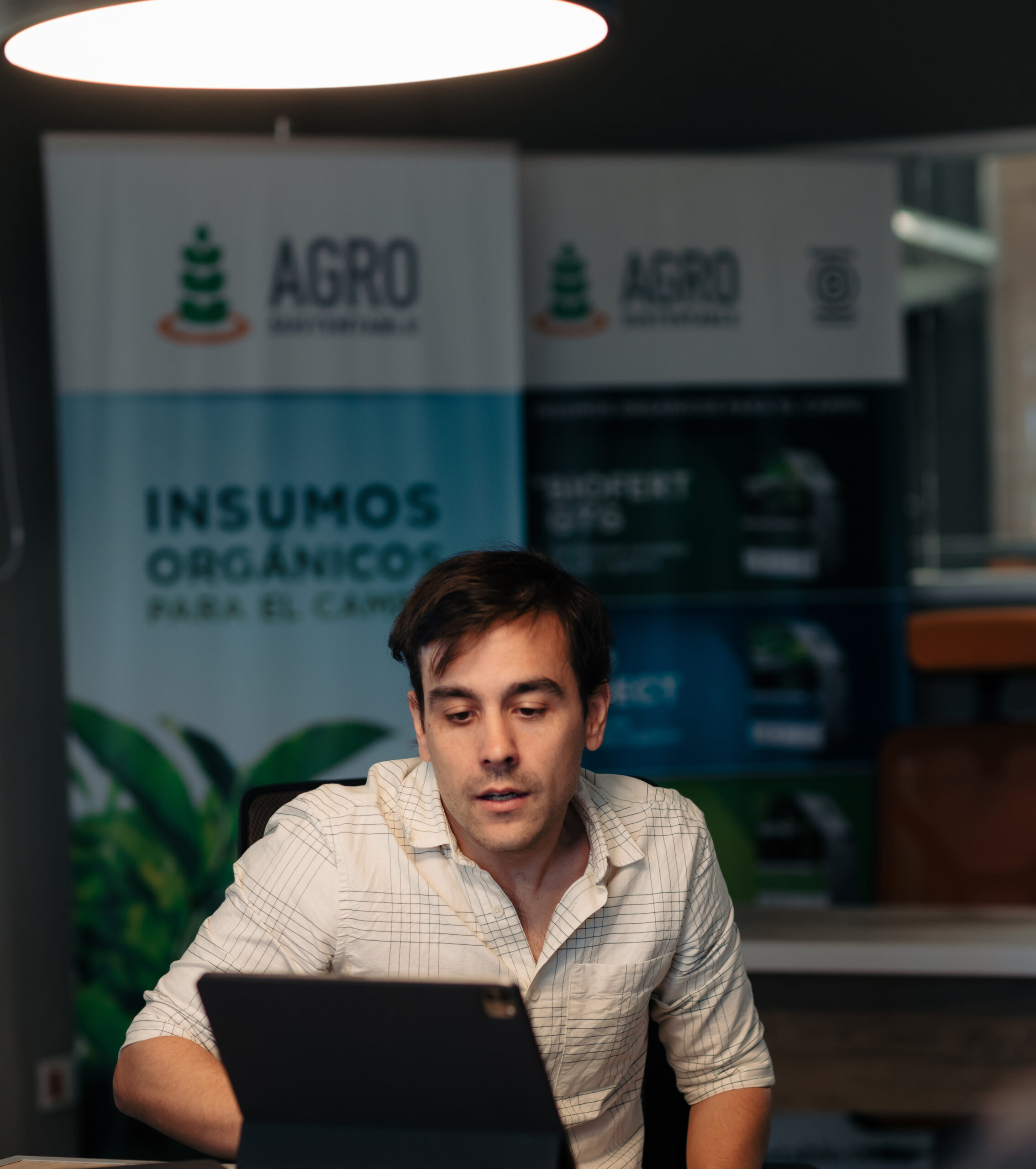 Matías Imperiale, ingeniero agrónomo, sentado frente a una notebook en las oficinas de Agro Sustentable, con carteles institucionales de fondo que promocionan insumos orgánicos para el campo.