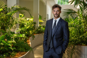 Hombre de traje oscuro posando en un entorno interior rodeado de plantas verdes y luz natural.