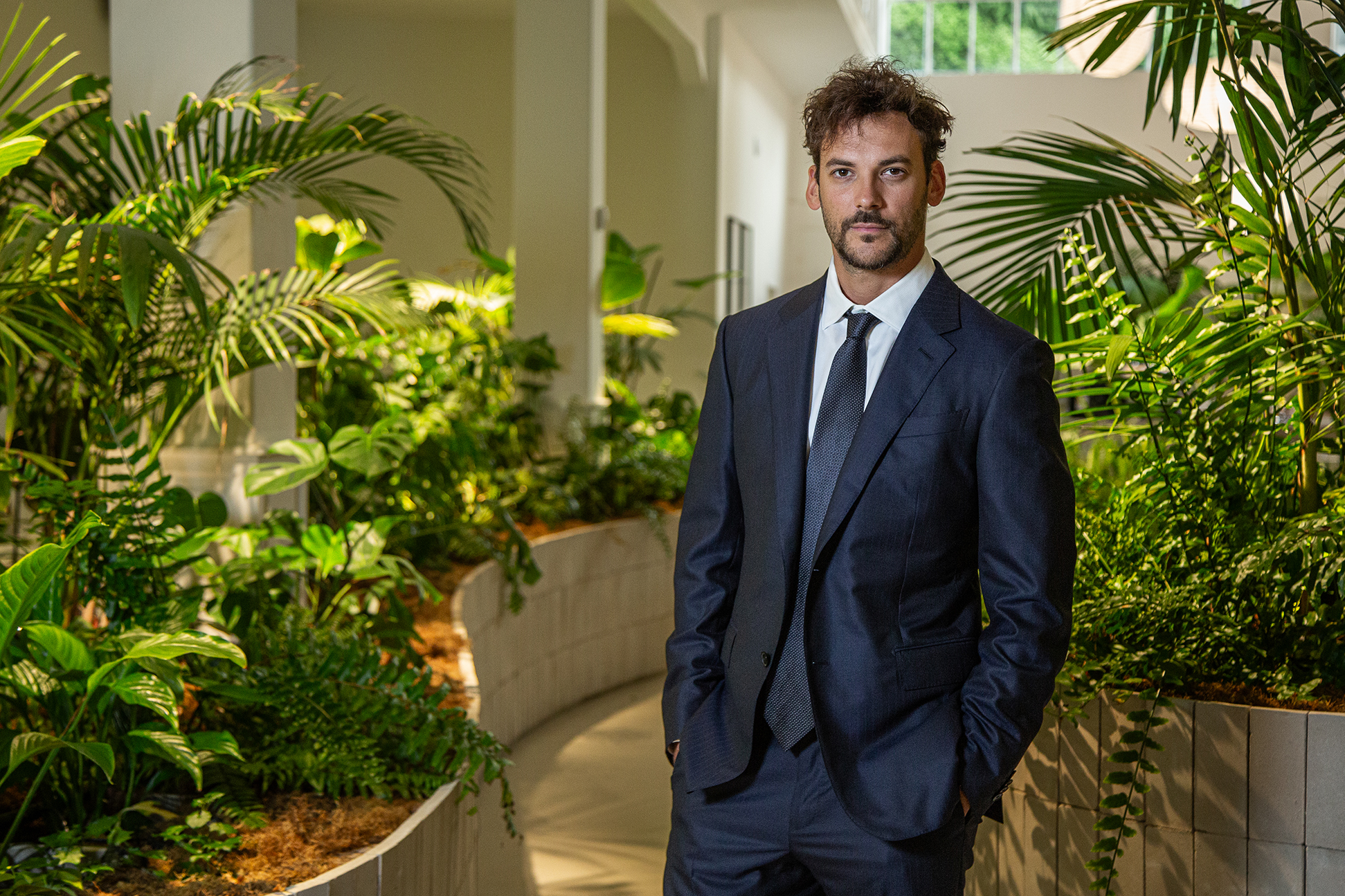 Hombre de traje oscuro posando en un entorno interior rodeado de plantas verdes y luz natural.