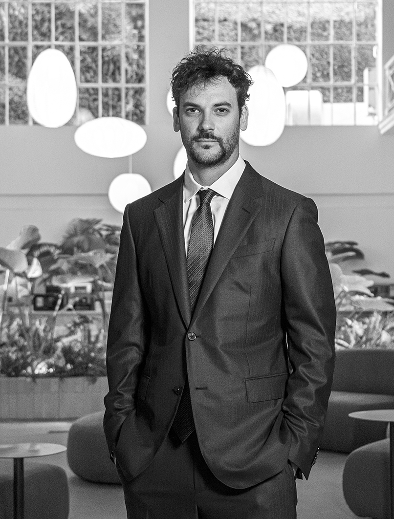 Hombre joven con traje formal en un entorno moderno y natural, posando con confianza en un espacio iluminado y elegante.