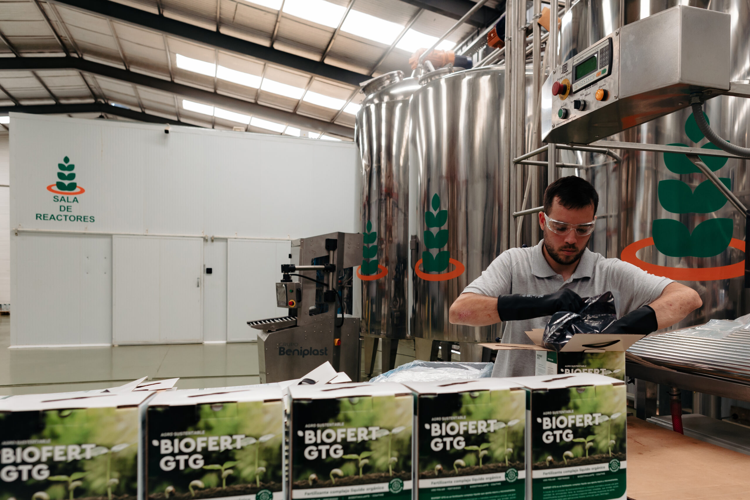 Trabajador de Agro Sustentable empacando cajas de biofertilizante orgánico BIOFERT GTG en una planta con tanques de acero y sala de reactores.