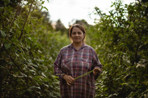 Mujer agricultora de pie entre hileras de cultivos verdes, sosteniendo una planta, rodeada de vegetación.